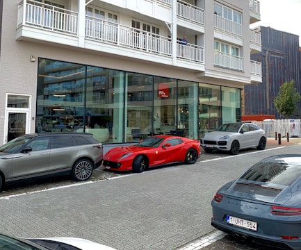 A street scene featuring several luxury cars parked in front of a modern building with large glass windows. The cars include a red sports car and two SUVs. The building has multiple balconies and a sign indicating it may be a residential place.