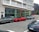 A street scene featuring several luxury cars parked in front of a modern building with large glass windows. The cars include a red sports car and two SUVs. The building has multiple balconies and a sign indicating it may be a residential place.