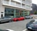 A street scene featuring several luxury cars parked in front of a modern building with large glass windows. The cars include a red sports car and two SUVs. The building has multiple balconies and a sign indicating it may be a residential place.