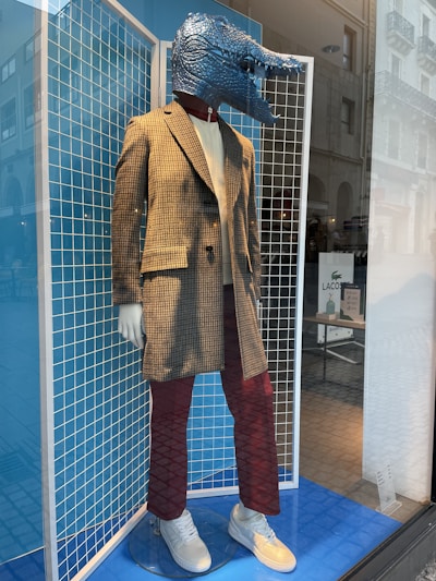 A mannequin dressed in stylish attire features a distinctive crocodile headpiece. It wears a plaid blazer over a light shirt, paired with maroon pants and white sneakers. The background consists of grid panels and a blue floor, set in a store display with exterior reflections visible on the glass.