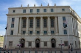 A large neoclassical building with multiple columns and tall windows dominates the scene. The facade is made of light-colored stone, and the building is identified as the United States Post Office and Court House. There are two flags on poles in front of the building, along with several lampposts and planters with flowers lining the pavement.