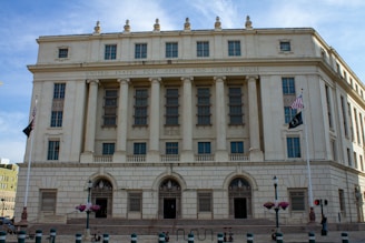 A large neoclassical building with multiple columns and tall windows dominates the scene. The facade is made of light-colored stone, and the building is identified as the United States Post Office and Court House. There are two flags on poles in front of the building, along with several lampposts and planters with flowers lining the pavement.