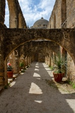 An architectural pathway features stone arches creating a sequence of shadows along a gravel path. The dome with a cross at the top is visible in the background. Potted plants, including a slender-leaved yucca and red flowers, line the sides of the pathway under the blue sky.