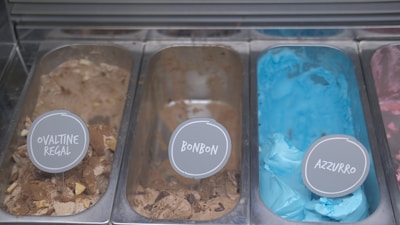 Three tubs of ice cream with distinct colors and labels are placed in a display freezer. The leftmost tub is filled with brown ice cream labeled 'Ovaltine Regal', the middle tub contains a lighter brown ice cream named 'Bonbon', and the rightmost tub showcases a bright blue colored ice cream called 'Azzurro'. Each tub has a small sign with the corresponding flavor name placed on top.