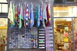 A variety of colorful flags hang above a brick facade, below which numerous postcards are displayed in neat rows. The scene is set outside a souvenir shop filled with various gifts, including items like magnets and postcards.