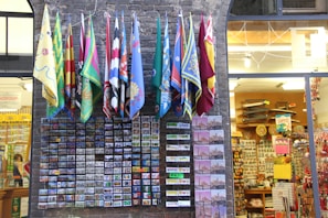 A variety of colorful flags hang above a brick facade, below which numerous postcards are displayed in neat rows. The scene is set outside a souvenir shop filled with various gifts, including items like magnets and postcards.