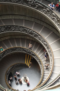 A spiral staircase with ornate designs on the railings, viewed from above. People are walking up and down the stairs, and some are standing on the landing below. The stairs are gray with occasional yellowish elements, and the landing below appears dark with a central circular feature.