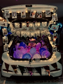 A car trunk is beautifully decorated for a birthday celebration. It features colorful balloons in shades of pink, purple, and blue, accompanied by string lights and garlands adding a festive touch. Hanging photographs and balloons shaped like stars are displayed around the edges. A prominent 'Happy Birthday' banner is centered among the decorations, creating a lively party atmosphere.