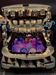 A car trunk is beautifully decorated for a birthday celebration. It features colorful balloons in shades of pink, purple, and blue, accompanied by string lights and garlands adding a festive touch. Hanging photographs and balloons shaped like stars are displayed around the edges. A prominent 'Happy Birthday' banner is centered among the decorations, creating a lively party atmosphere.