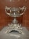 A highly detailed and ornate silver trophy or cup sits on display. It features intricate engravings and elaborate handles with decorative embellishments. The base of the cup includes detailed sculptures of mythical creatures or ornate designs.