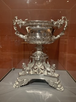 A highly detailed and ornate silver trophy or cup sits on display. It features intricate engravings and elaborate handles with decorative embellishments. The base of the cup includes detailed sculptures of mythical creatures or ornate designs.