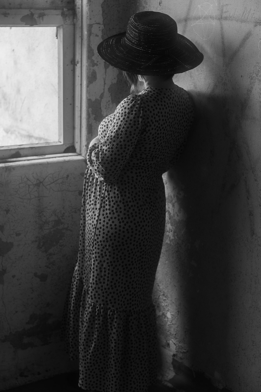 a woman in a dress and hat leaning against a wall