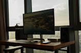 A modern workspace featuring a large computer monitor displaying a mountainous landscape. The setup is placed on a wooden desk with speakers on either side. A keyboard and mouse are neatly positioned in front of the monitor, and additional technological devices are placed nearby. The setup is situated in a room with large windows, allowing natural light to create a serene atmosphere.