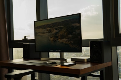 A modern workspace featuring a large computer monitor displaying a mountainous landscape. The setup is placed on a wooden desk with speakers on either side. A keyboard and mouse are neatly positioned in front of the monitor, and additional technological devices are placed nearby. The setup is situated in a room with large windows, allowing natural light to create a serene atmosphere.