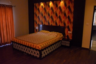 A bedroom features a large bed with a colorful patterned bedspread against a wall of geometric diamond patterns in shades of orange, red, and brown. The floor is dark wood, and there is a window with orange curtains to the left. Soft lighting creates a warm and cozy ambiance.