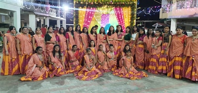 A group of women and girls dressed in traditional, colorful attire gather in front of a decorated backdrop with bright lights. The background features vibrant yellow and pink curtains adorned with floral arrangements and string lights, suggesting a festive or ceremonial event. The ground is paved, and the setting appears to be an outdoor space at night.