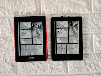 Two Kindle devices are mounted on a textured white wall. Each device displays the same page from a black-and-white manga. The manga panels depict characters having a conversation with some text bubbles containing the words 'Devil' and 'If you're gonna kill me, do it. I'm dead either way.' The Kindle on the left has a red case, while the one on the right has a black case.