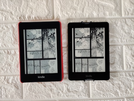 Two Kindle devices are mounted on a textured white wall. Each device displays the same page from a black-and-white manga. The manga panels depict characters having a conversation with some text bubbles containing the words 'Devil' and 'If you're gonna kill me, do it. I'm dead either way.' The Kindle on the left has a red case, while the one on the right has a black case.