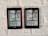 Two Kindle devices are mounted on a textured white wall. Each device displays the same page from a black-and-white manga. The manga panels depict characters having a conversation with some text bubbles containing the words 'Devil' and 'If you're gonna kill me, do it. I'm dead either way.' The Kindle on the left has a red case, while the one on the right has a black case.