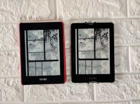 Two Kindle devices are mounted on a textured white wall. Each device displays the same page from a black-and-white manga. The manga panels depict characters having a conversation with some text bubbles containing the words 'Devil' and 'If you're gonna kill me, do it. I'm dead either way.' The Kindle on the left has a red case, while the one on the right has a black case.