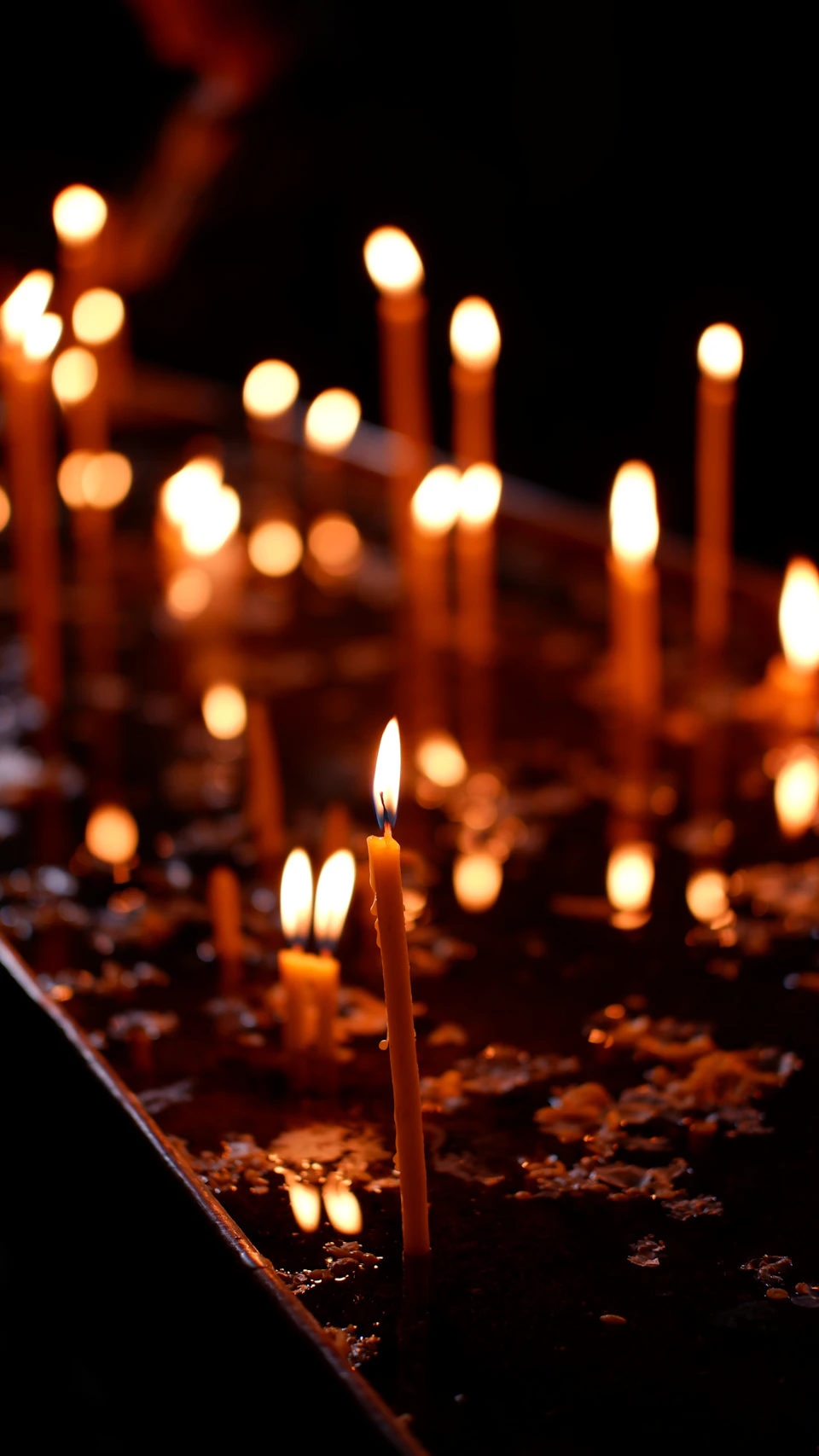 The image shows a group of lit candles arranged on a dark surface, possibly a table or a countertop. The candles are positioned at various angles and heights, creating an interesting visual effect with their reflections and shadows. Some of the candles appear to be burning brightly, while others seem to have just been lit, casting a warm glow throughout the scene.