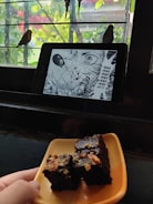 A tablet displaying a manga page with an intense scene is placed on a window ledge. Outside, two birds are perched on the bars of the window. In the foreground, a hand is holding a plate with pieces of chocolate cake topped with nuts.