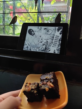 A tablet displaying a manga page with an intense scene is placed on a window ledge. Outside, two birds are perched on the bars of the window. In the foreground, a hand is holding a plate with pieces of chocolate cake topped with nuts.