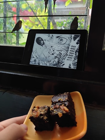 A tablet displaying a manga page with an intense scene is placed on a window ledge. Outside, two birds are perched on the bars of the window. In the foreground, a hand is holding a plate with pieces of chocolate cake topped with nuts.
