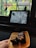 A tablet displaying a manga page with an intense scene is placed on a window ledge. Outside, two birds are perched on the bars of the window. In the foreground, a hand is holding a plate with pieces of chocolate cake topped with nuts.