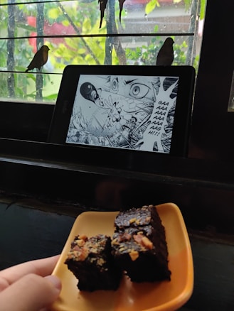 A tablet displaying a manga page with an intense scene is placed on a window ledge. Outside, two birds are perched on the bars of the window. In the foreground, a hand is holding a plate with pieces of chocolate cake topped with nuts.