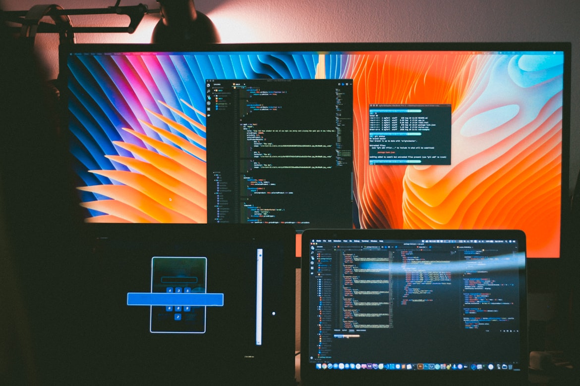 multi-screen setup displaying lines of code in one of them, a bright orange-blue abstract image on another