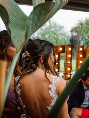 A woman with her hair styled in an elegant updo adorned with small flowers, wearing a lace dress with an open back. The foreground features large green leaves, and in the background, there are illuminated marquee lights. Another person is partially visible next to her, seated in a formal setting.