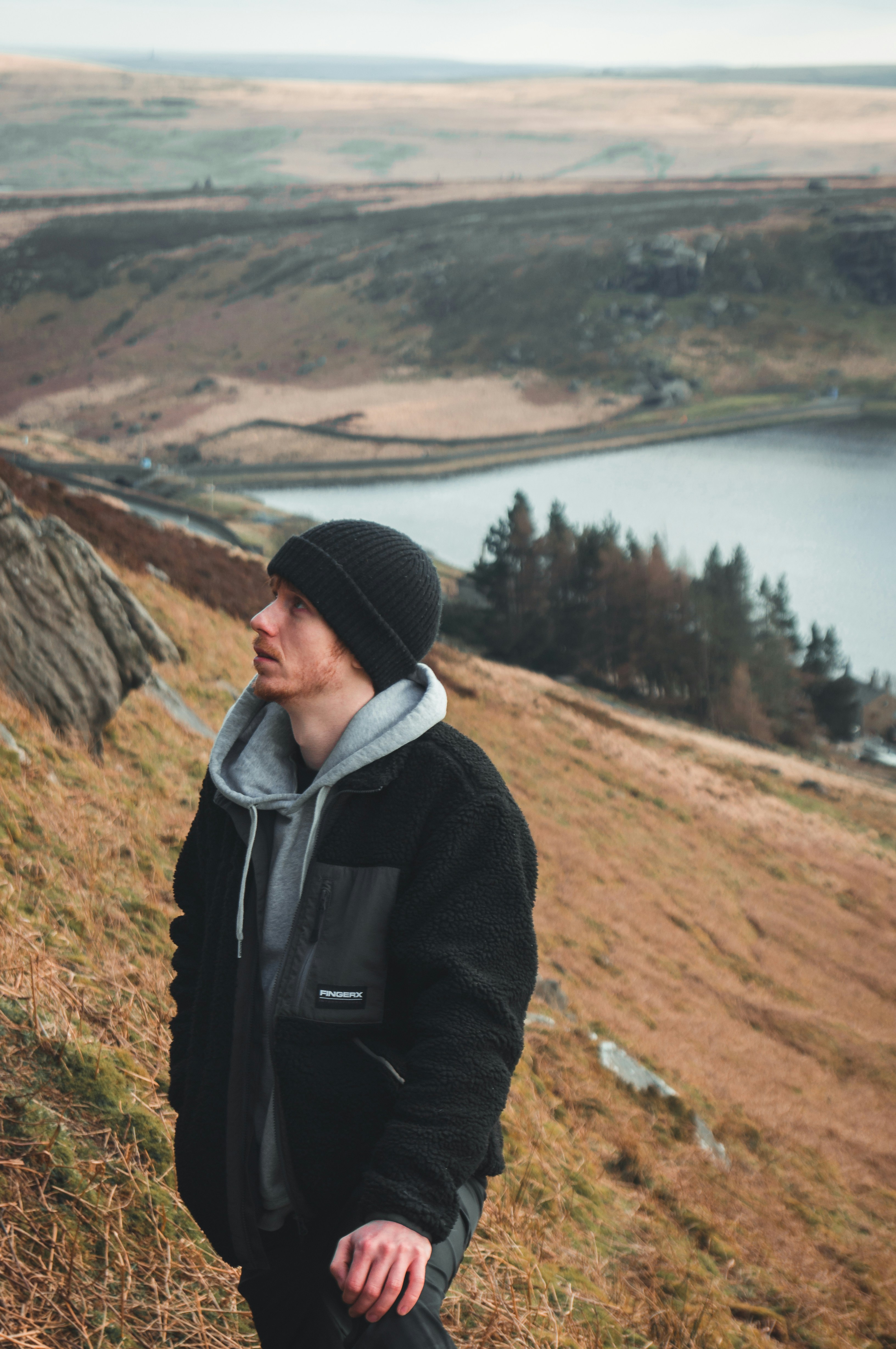 A man in a dark jacket and a hat looks into the distance, with hills and a reservoir in the background.