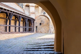 A narrow cobblestone alleyway is flanked by old stone buildings with wooden supports. An archway provides a view into a secluded courtyard featuring a vintage street lamp attached to the wall. The warm tones of the stone and wood create a rustic and historic atmosphere.