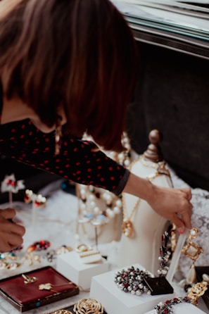 A person is reaching out towards various pieces of jewelry displayed on a table. The collection includes necklaces, rings, and earrings, arranged on stands and boxes. The person is wearing a dark, polka-dotted shirt, and their hand is interacting with the jewelry, possibly arranging or selecting an item.