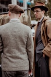Two men are engaged in conversation, wearing vintage-inspired clothing. One man, facing away from the camera, is in a brown beret and gray tweed jacket. The other, wearing glasses and a brown hat, is dressed in a camel-colored overcoat over a blue suit.