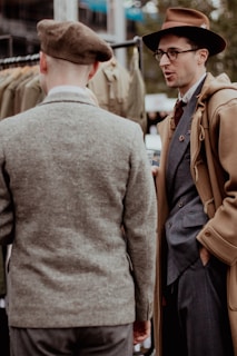 Two men are engaged in conversation, wearing vintage-inspired clothing. One man, facing away from the camera, is in a brown beret and gray tweed jacket. The other, wearing glasses and a brown hat, is dressed in a camel-colored overcoat over a blue suit.