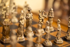 Intricately designed chess pieces are displayed on a wooden chessboard. The pieces are metallic, with a focus on a prominent king figure at the center, surrounded by other finely detailed pieces. The shallow depth of field creates a blurred background effect, highlighting the central chess piece.