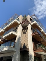 A modern multi-story building with large balconies and glass railings, featuring a blend of stone and wood materials. A Buddha statue is positioned in a central alcove. The building is surrounded by clear blue skies and a few clouds.