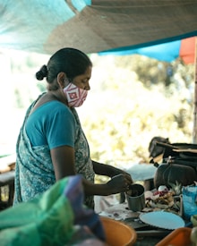 A woman wearing a patterned face mask and a floral sari is preparing food at a makeshift outdoor kitchen. She is focused on her task, holding a stainless steel cup. Various ingredients are scattered on the table along with kitchen utensils. The setting has a calm atmosphere, with diffused natural light visible under the tarp above.