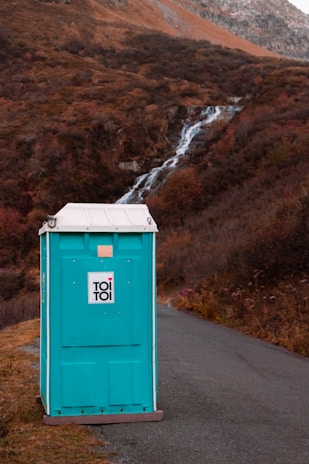 A blue portable toilet stands on a paved pathway surrounded by natural landscape. In the background, a small waterfall flows down a rocky hillside covered in autumn foliage.