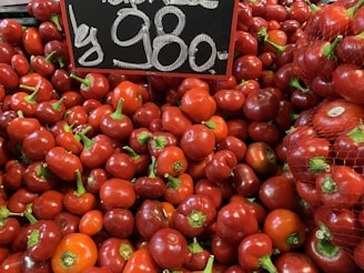 A large pile of red bell peppers is displayed, with some still in a mesh bag. A chalkboard sign above them shows the price in large white numbers.