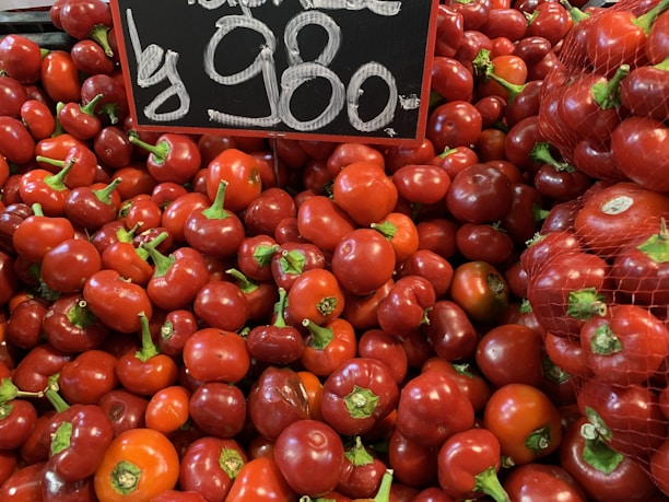A large pile of red bell peppers is displayed, with some still in a mesh bag. A chalkboard sign above them shows the price in large white numbers.