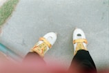 A pair of roller skates with a white base and orange-yellow accents worn by a person standing on a gray concrete surface. Part of the person's leg is visible, dressed in black clothing. There is a green rail and a patch of grass in the corner of the image.