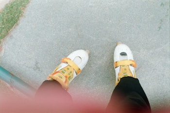 A pair of roller skates with a white base and orange-yellow accents worn by a person standing on a gray concrete surface. Part of the person's leg is visible, dressed in black clothing. There is a green rail and a patch of grass in the corner of the image.