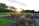 A landscaped garden with flower beds and colorful blooms, bordered by stone edging and winding pathways. The sun is setting in the background, casting a warm glow over the area. Trees with lush foliage line the perimeter, adding to the serene and natural atmosphere.