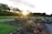 A landscaped garden with flower beds and colorful blooms, bordered by stone edging and winding pathways. The sun is setting in the background, casting a warm glow over the area. Trees with lush foliage line the perimeter, adding to the serene and natural atmosphere.
