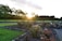 A landscaped garden with flower beds and colorful blooms, bordered by stone edging and winding pathways. The sun is setting in the background, casting a warm glow over the area. Trees with lush foliage line the perimeter, adding to the serene and natural atmosphere.