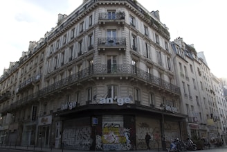 A classic Parisian building with ornate wrought iron balconies and white shutters on multiple floors. The street-level facade is covered in graffiti, and there are bicycles parked at the corner. A person is walking by, adding a sense of urban life to the scene.