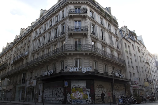 A classic Parisian building with ornate wrought iron balconies and white shutters on multiple floors. The street-level facade is covered in graffiti, and there are bicycles parked at the corner. A person is walking by, adding a sense of urban life to the scene.